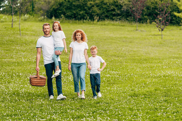Fototapeta premium happy family smiling at camera while walking with picnic basket in park