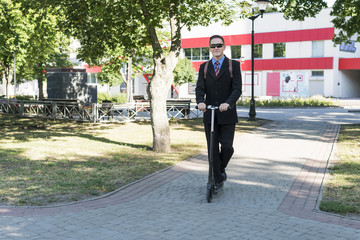 Businessman in sunglasses with a backpack riding down the street on a kick scooter