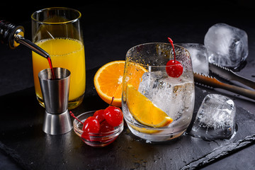 Cocktail concept. Cocktail making. Grenadine is poured into a jigger. Maraschino cherry, ice cube and orange slice in a glass on a black slate tray.