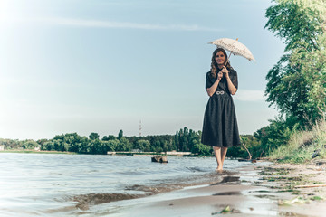 a woman walks along the river's edge hiding from the sun under lace umbrella