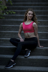 Fototapeta premium Young pretty sportive girl is sitting on the steps on the late summer evening looking down smiling