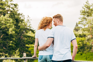 back view of redhead couple embracing and smiling each other while standing together in park