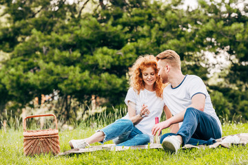 happy couple drinking wine while sitting on plaid at picnic in park