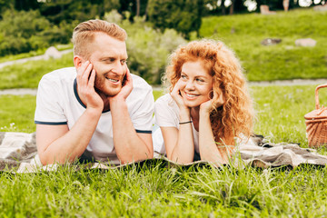 happy redhead couple smiling each other while lying together on plaid at picnic