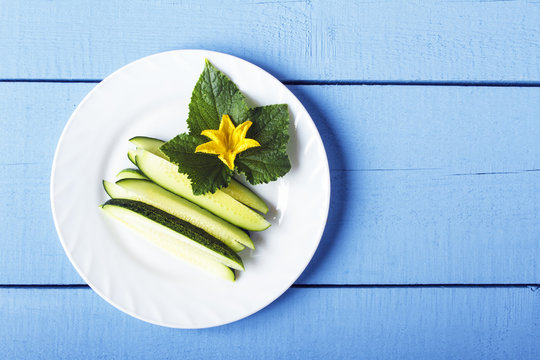 Freshly sliced cucumbers, green leaves and yellow flowers on white plate. Healthy food concept. Top view on wooden table. Copy space