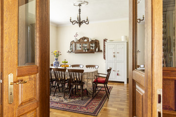 Real photo of an antique dining room interior with a big table, chairs, wall cupboard and carpet. View through a door