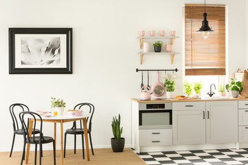 Painting in a black frame in a dining room interior with a table and chairs next to a kitchen interior with tools, plants and cupboards, Real photo - obrazy, fototapety, plakaty