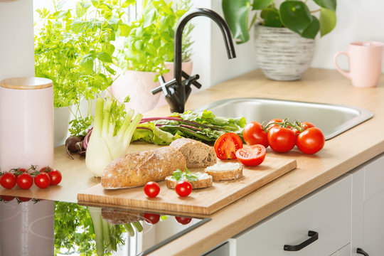 Herbs, bread and tomatoes on a cutting board next to a sink in a kitchen interior