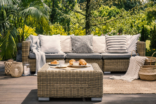 Close-up of a rattan outdoor table with coffee and croissants on it in front of a cozy sofa with white pillows on the patio of a spa hotel