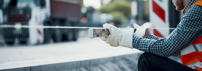 Road worker leveling a string line, holding the tool with safety gloves and a blurry construction site in the background