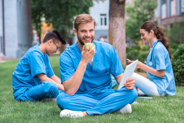 Naklejka premium handsome caucasian medical student eating apple and holding tablet