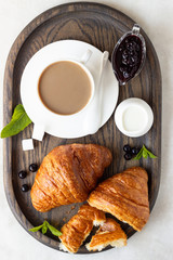 Breakfast concept: two fresh french croissant with a cup of coffee and jam on a dark wooden tray.