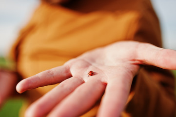 Stylish man in glasses, brown jacket and hat posed on green field and holding at hand ladybug.
