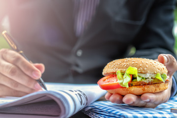 Businessman holding hamburger in one hand while holding pen in another hand pointing at newspaper on table