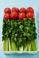 close up view of food container full of fresh tomatoes, parsley and celery on blue background