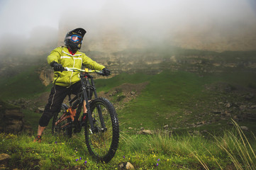 Fototapeta premium An adult mtb cyclist on a mountain bike at the foot of a cliff surrounded by green grass. Low clouds. North Caucasus. Russia