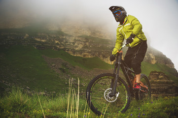 Fototapeta na wymiar An adult mtb cyclist on a mountain bike at the foot of a cliff surrounded by green grass. Low clouds. North Caucasus. Russia