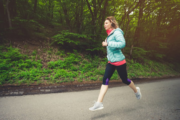 Young fitness blonde woman in headphones running at morning caucasian forest trail in sun light