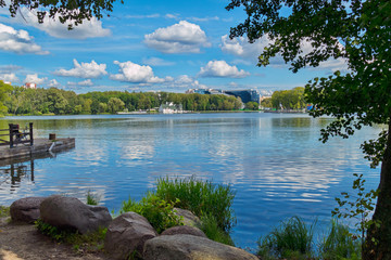 A delightful view of the large lake from the shore with large stones lying on it. With a blue sky over it with slowly sailing white bales.