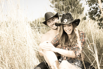 Cowboy and cowgirl couple sitting in grass, laughing as they tickle each other.