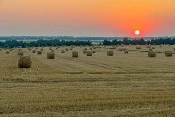 A lot of stacks of hay on the field under the sun setting in the horizon is red