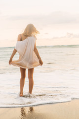 Fototapeta na wymiar Hipster trendy girl hold her long casual summer dress and walk in waves on the coastline. Sporty lady on sea sand beach sunset or ocean sunrise. Travel, active, yoga, freedom lifestyle concept.