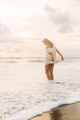 Hipster trendy girl hold her casual summer dress and walk in little waves on the coastline. Sporty lady on sea sand beach sunset or ocean sunrise. Travel, active, yoga lifestyle concept.