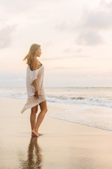 Hipster trendy girl in casual summer dress walk barefoot by the waterline and look to the little waves. Sporty lady on sea sand beach sunset or ocean sunrise. Travel, active, yoga lifestyle concept.