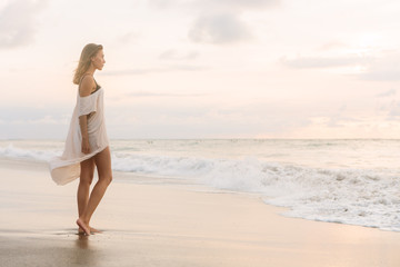 Hipster trendy woman in casual summer dress walking barefoot by the waterline and look to the waves. Sporty lady on sea sand beach sunset or ocean sunrise. Travel, active, yoga lifestyle concept.