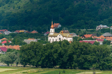 Fototapeta na wymiar Church with white walls and bulbs of domes with nearby village houses among the trees in a picturesque area on the background of a mountain slope.