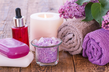 Fototapeta na wymiar Soap, red bottle with aromatic oil, burning candle, bowl with sea salt, lilac flowers and towels on wooden boards