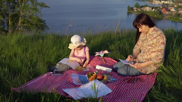 The family sits on a blanket on a picnic on a summer day on a hill overlooking a river or lake. Young mother and little daughter draw.
