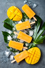 Mango popsicles over green tropical palm leaf on grey stone background. Overhead, top view
