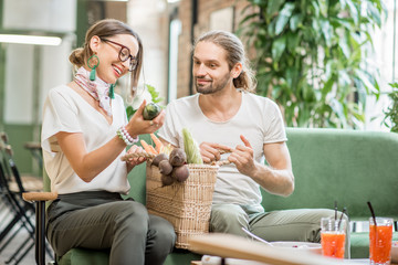 Young vegetarian couple sitting on the sofa with bag full of fresh vegetables in the beautiful green home interior