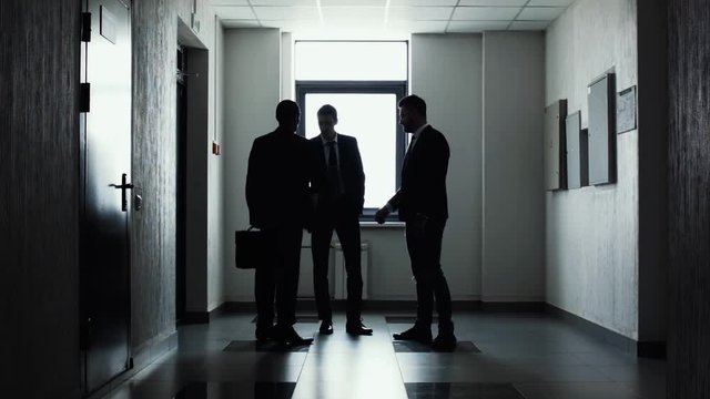 Silhouettes of three businessmen standing in corridor in front of light window. The end of working day. Team completed the work on the project. Businessmen say good-bye and shake hands. Handshake.