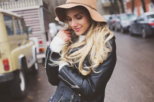 lifestyle portrait of young beautiful woman walking outdoor on city streets, wearing hat and leather jacket. Real people, candid shot.