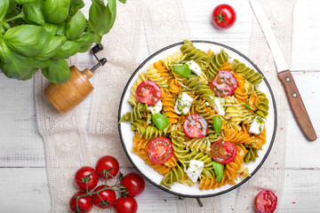 Italian food - Salad with colorful pasta, cherry tomatoes, feta cheese and fresh basil on white wooden background
