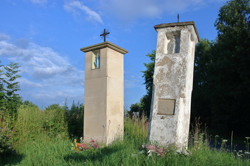 Dwie murowane  kapliczki przydrożne, jedna zabytkowa, stara, zniszczona, pochylona, obok niej nowa, stoi prosto, w tle zieleń, błękitne niebo