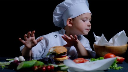 Cute boy is cooking a burger, wearing chefs suit, surrounded with food ingredintes on the black background