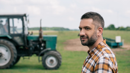side view of handsome middle aged farmer smiling while standing near tractor in field