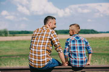 back view of father and son sitting on fence and smiling each other at farm