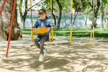 A girl on a walk reading a book swinging on a swing at the playground