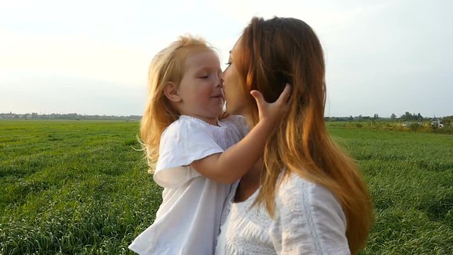 Happy family, mother and her little daughter walk on the green grass, summer evening. Mom with kid walking on green fields. Daughter caressing and kissing her mom. Slow motion