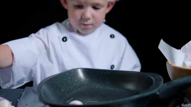 Cute boy is cooking a dish, wearing chefs suit and cap. Black background for commercial