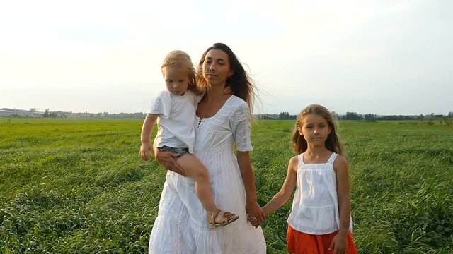 Happy family, mother and her little daughters walk on the green grass, summer evening. Mom with children walking on green fields. Slow motion