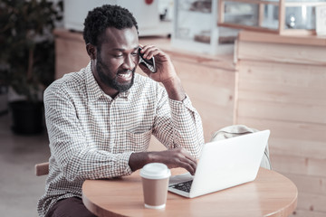 In the cafeteria. Delighted positive afro American man working on a netbook and making a call while...