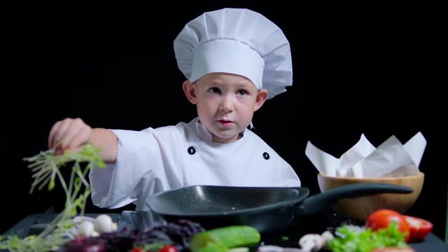 Cute boy is cooking a dish, wearing chefs suit and cap. Black background for commercial