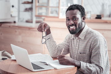 Positive mood. Happy cheerful afro American man sitting at the table and looking at you while enjoying his work