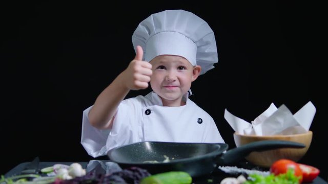 Cheerful boy shows thumb while cooking. He wears chefs suit and cap. Black background