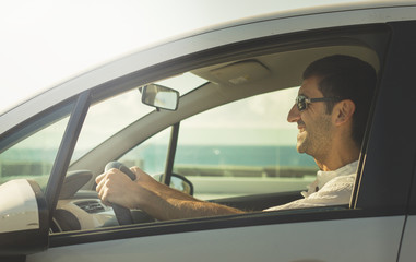 Happy man driving white car on sunny day by the coast. Smiling young driver with sunglasses and white shirt. Road trip, auto rental, insurance concepts